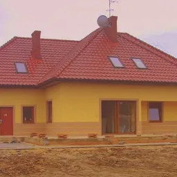 Żółty dom jednorodzinny z czerwoną dachówką, widoczne okna dachowe, komin i antena satelitarna, elewacja z poziomym pasem w innym odcieniu, teren wokół domu w trakcie prac wykończeniowych.